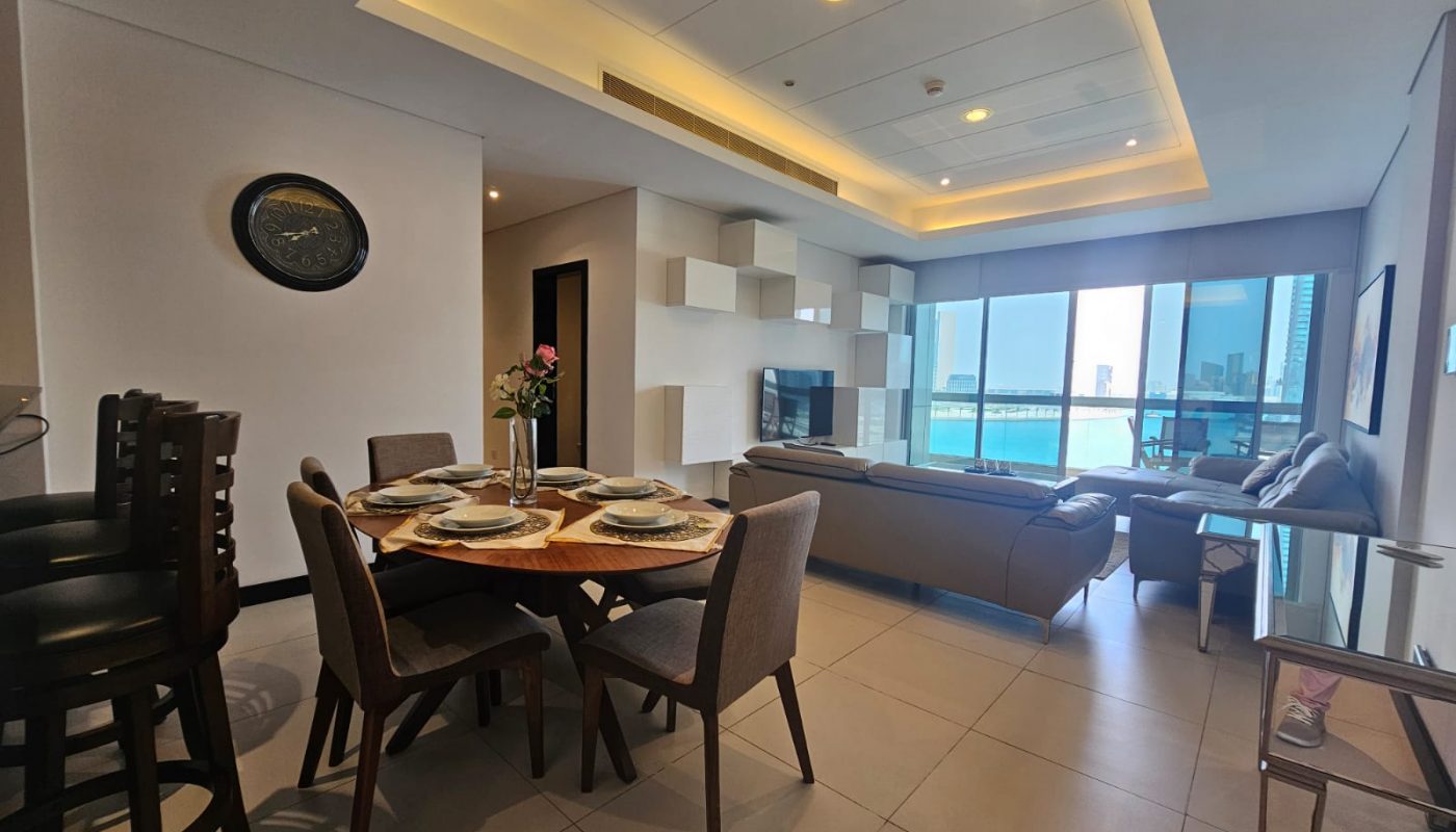 Modern open-plan living and dining area with a round table set for four, a sofa, large windows with a city and water view, and neutral décor.