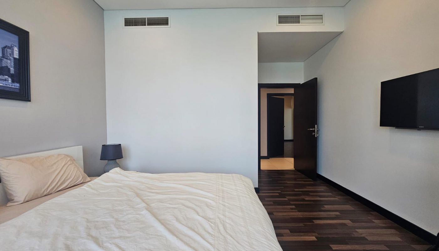 A modern bedroom with a bed, lamp, wall-mounted TV, wooden floor, and an open door leading to another room.