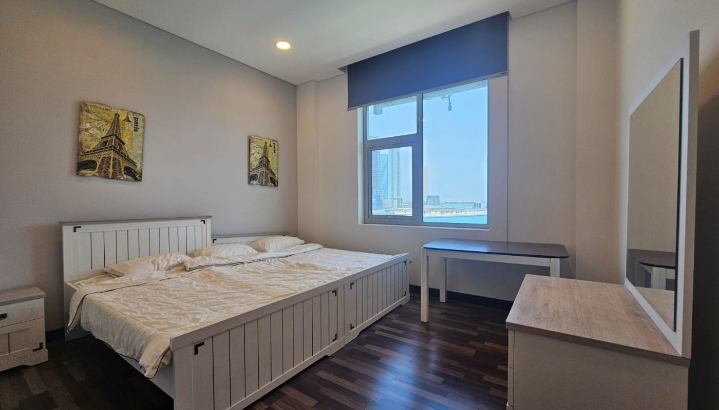 A bright bedroom with a double bed, white furniture, wall art, a window with city views, and dark wood flooring.