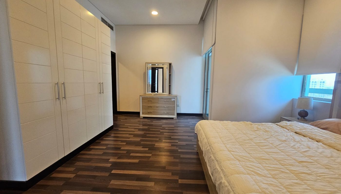 A bedroom with a double bed, white wardrobe, dresser with mirror, nightstand, lamp, and window with blinds, featuring wooden flooring and neutral walls.