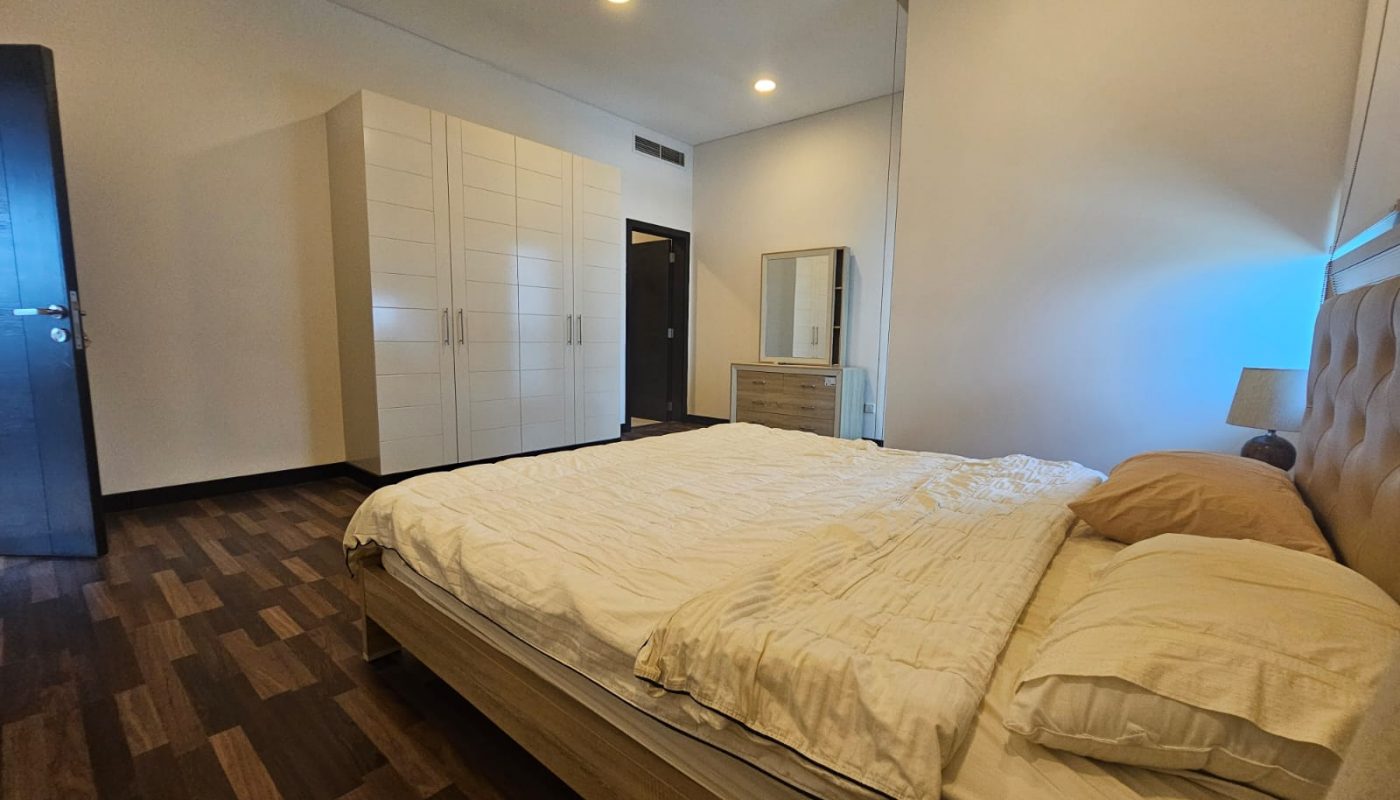 A bedroom with a double bed, wooden floor, white wardrobe, dresser with a mirror, bedside table, and neutral-colored walls.