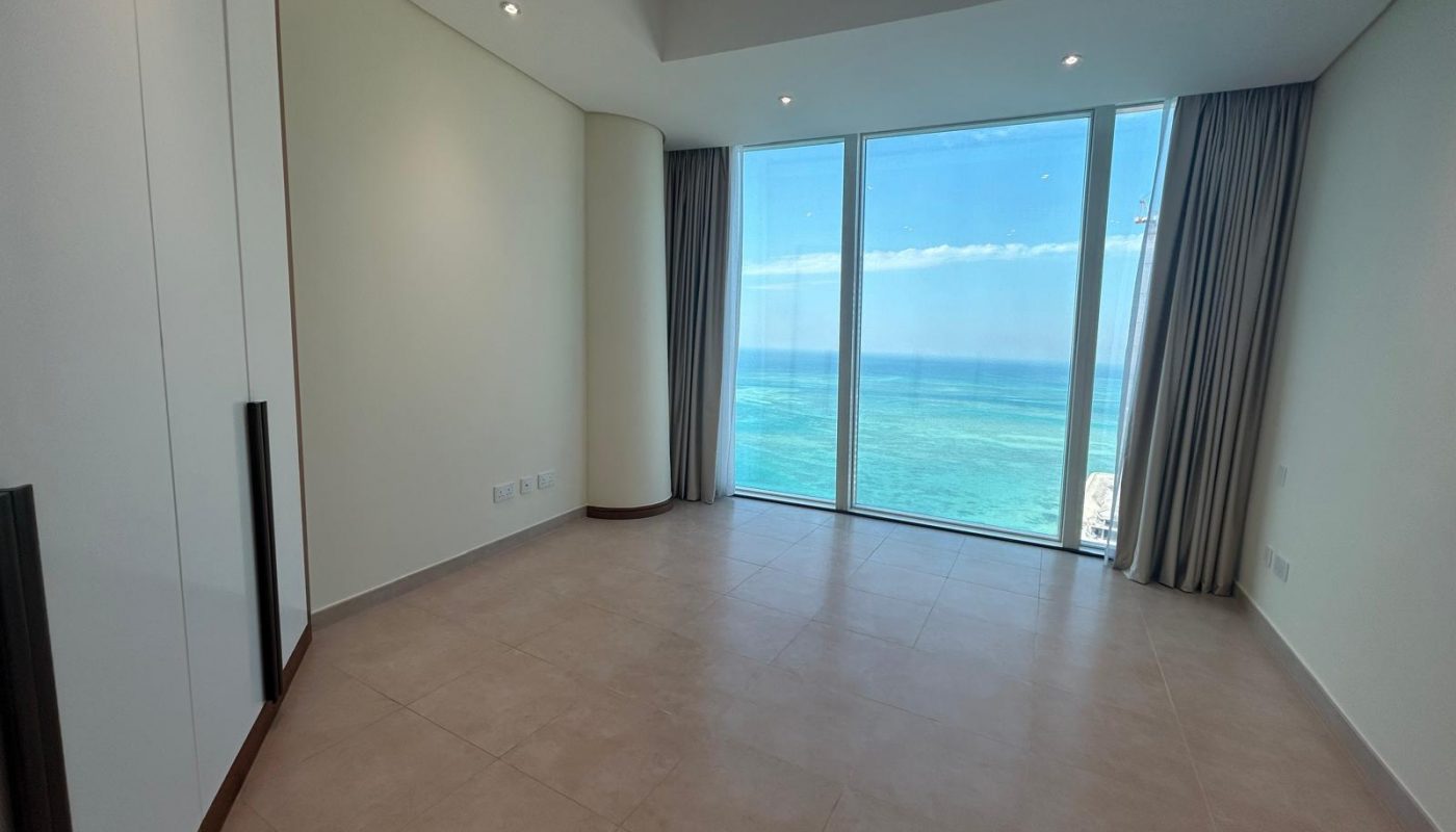 Empty room with tiled floor, white walls, built-in wardrobe, and large floor-to-ceiling window offering a view of the ocean and sky.