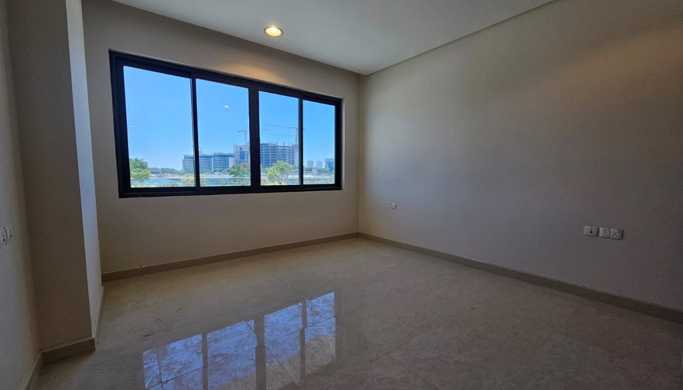 Empty, unfurnished room with beige tiled floor, white walls, and a large window showing an urban view with buildings and trees under a clear blue sky.