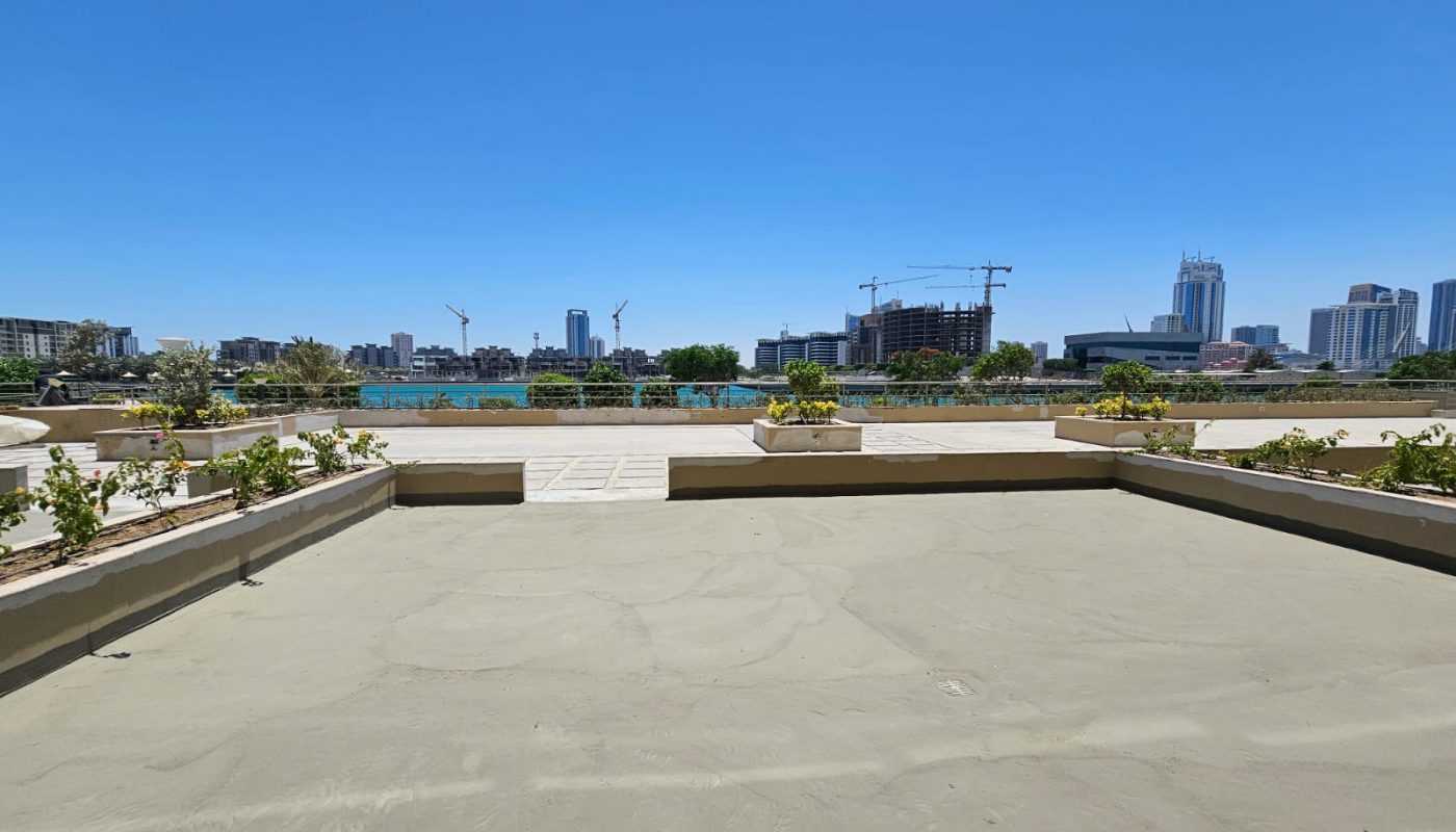 View of a city skyline with several buildings and cranes, seen from a rooftop terrace with planters and paved areas under a clear blue sky.