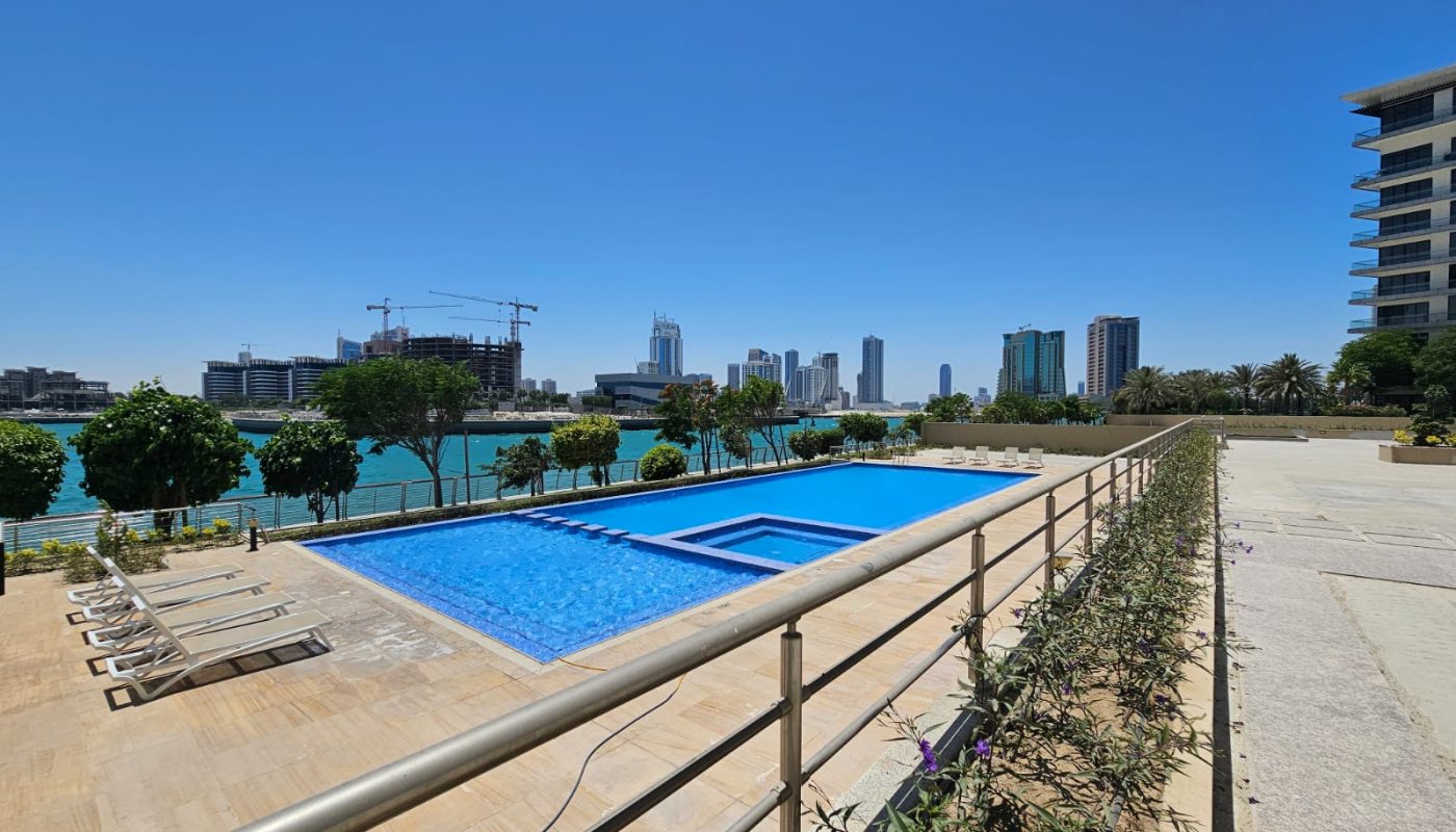 Outdoor swimming pool with lounge chairs beside a waterfront, city skyline and construction cranes visible in the background under a clear blue sky.