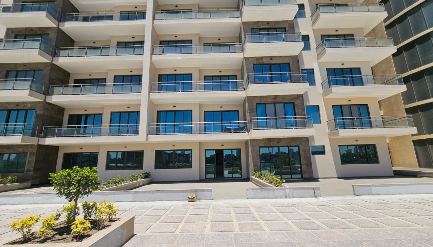 A modern multi-story apartment building with balconies, large glass windows, and landscaped planters in front.