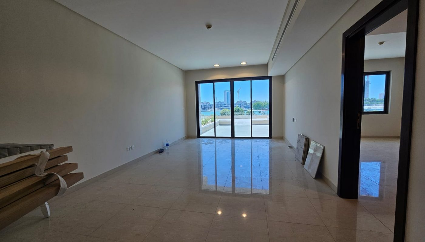 Unfurnished, empty room with tiled floor, large window doors leading to a balcony, and a doorway to an adjacent room. Some rolled carpets and frames rest against the walls.