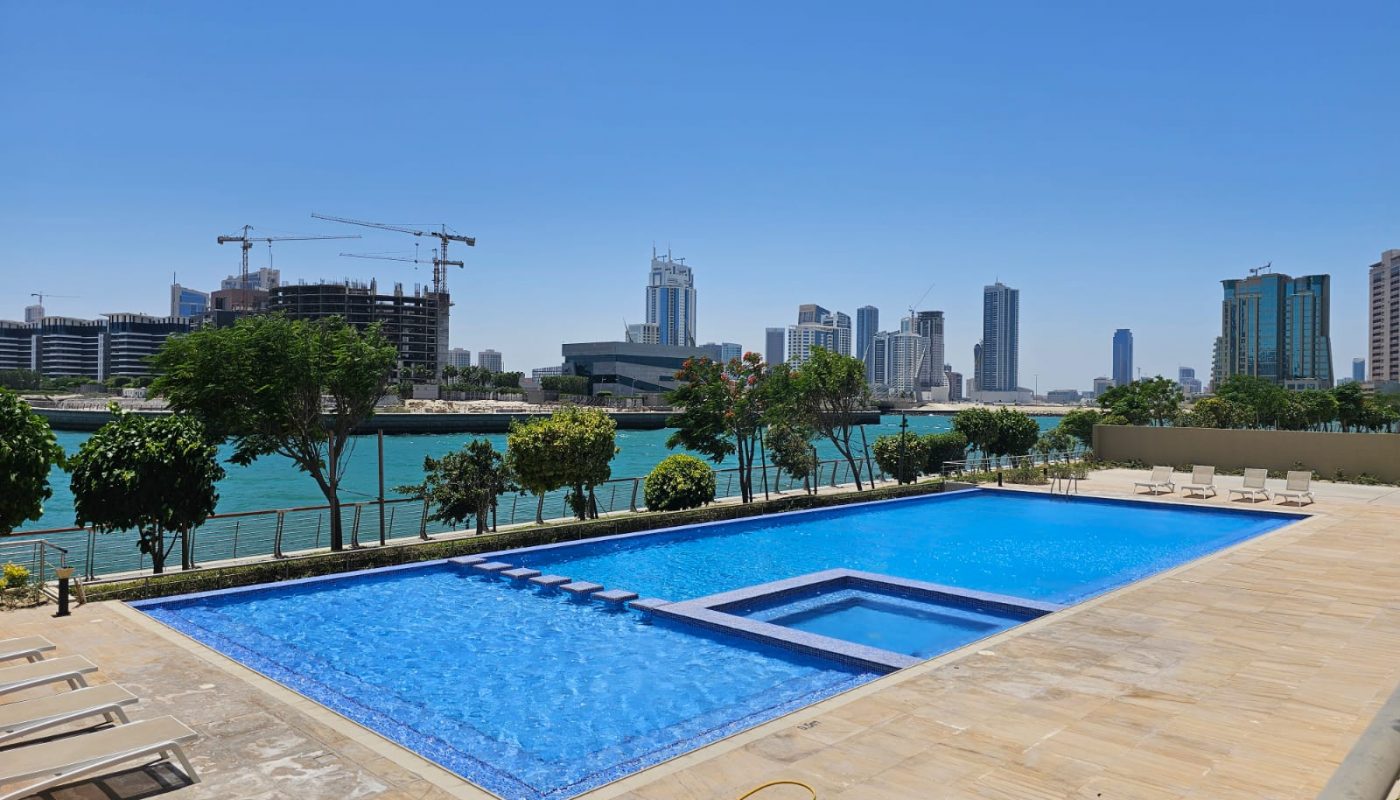 Outdoor swimming pool with a smaller connected pool, surrounded by lounge chairs, overlooking a river with city buildings and construction cranes in the background under a clear blue sky.
