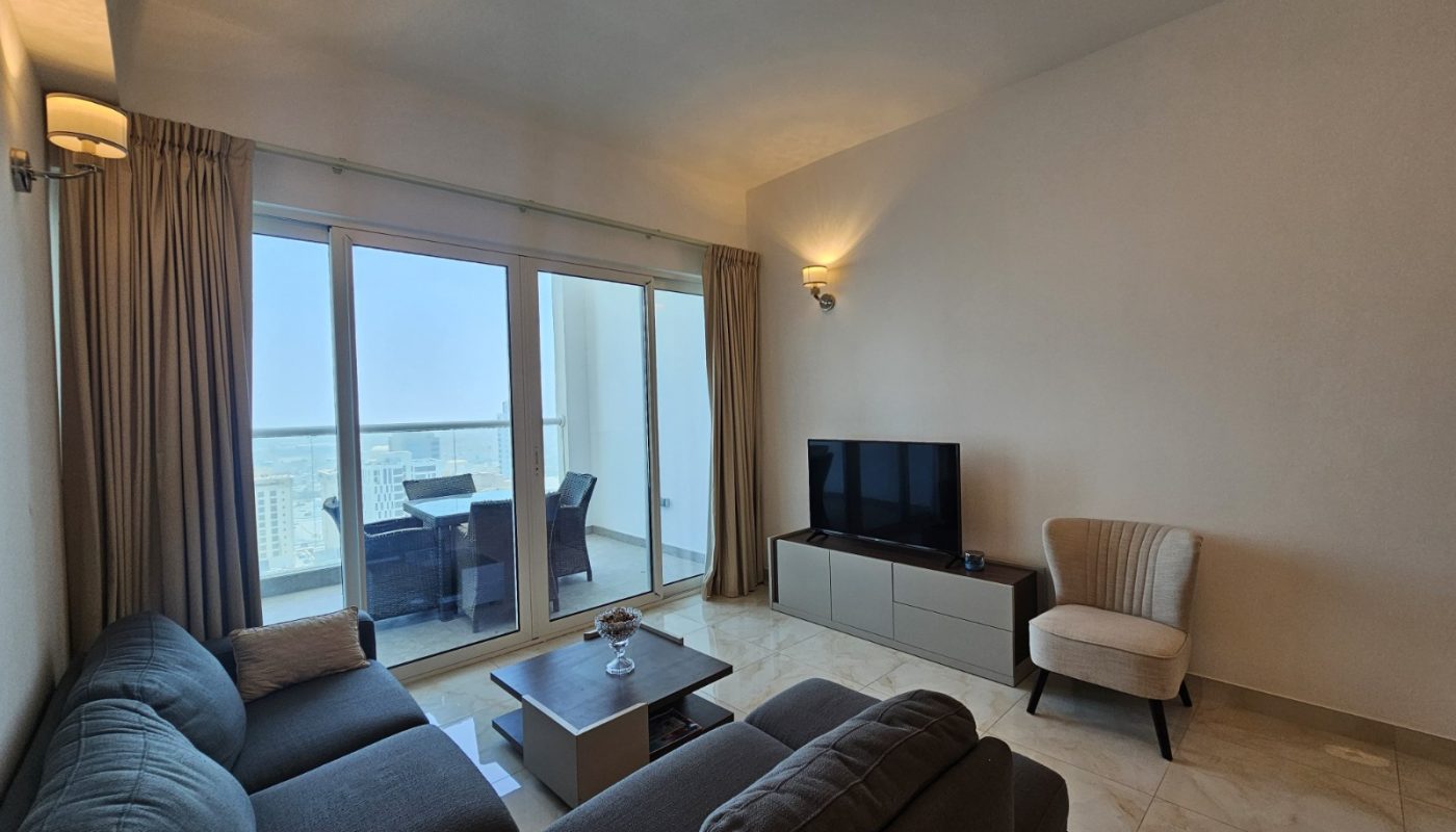 Modern living room with a grey sectional sofa, an armchair, a TV on a cabinet, a coffee table, and glass doors leading to a balcony with outdoor seating and a city view.