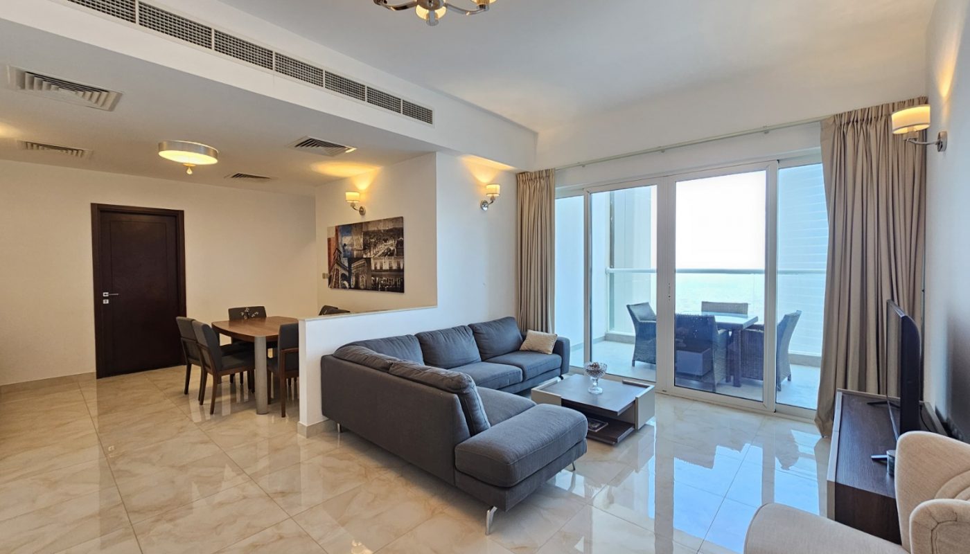 Modern living room with gray sectional sofa, dining table, chandelier lighting, tiled floor, wall art, large window, and balcony with outdoor seating.