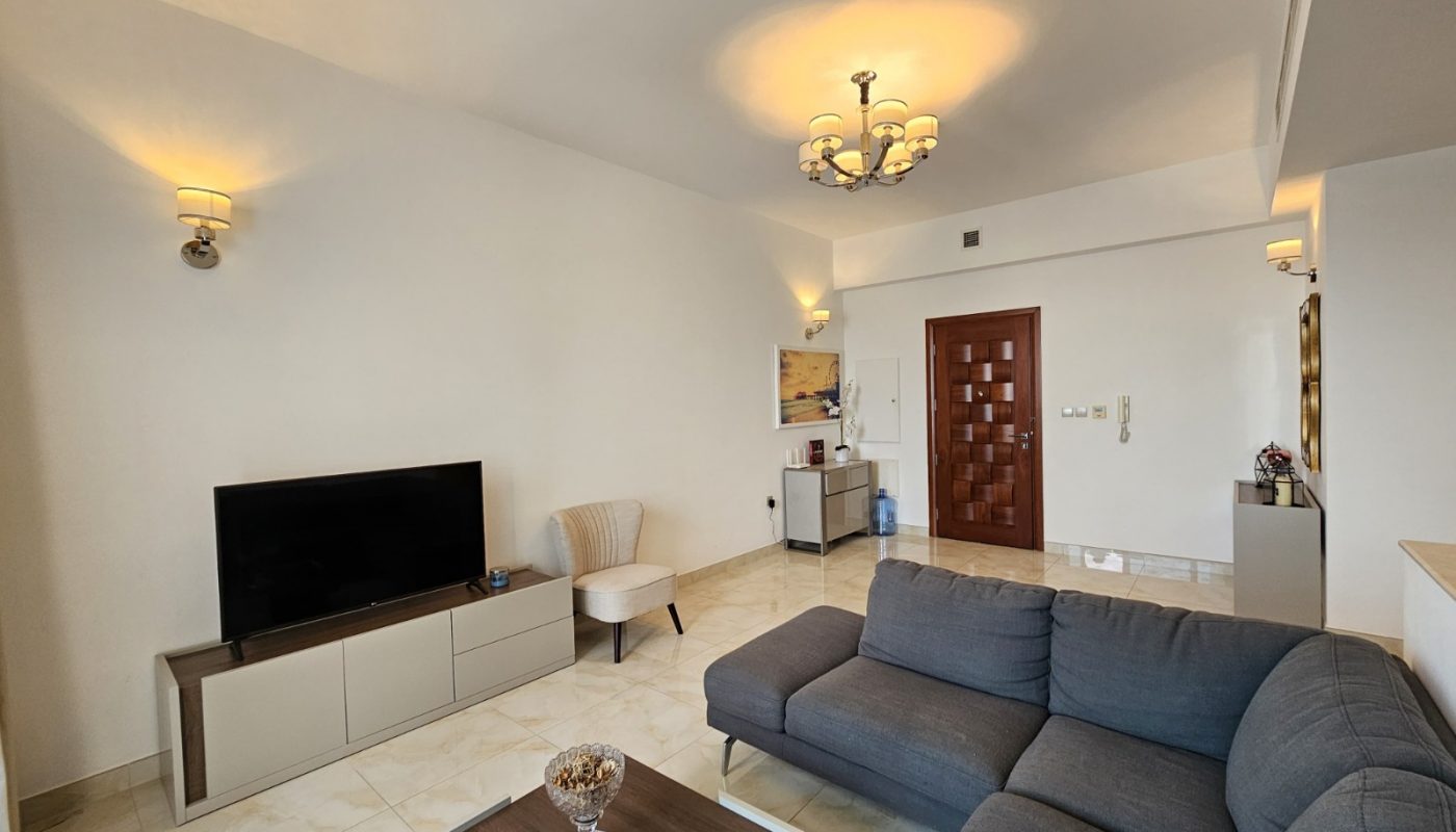 A modern living room with a gray sectional sofa, wall-mounted TV, small table, beige chair, overhead lighting, and neutral decor.
