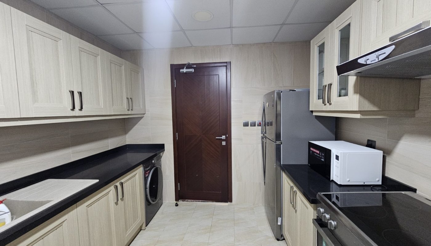 Modern kitchen with light wood cabinets, black countertops, stainless steel appliances, microwave, washing machine, and a closed dark brown door at the center.