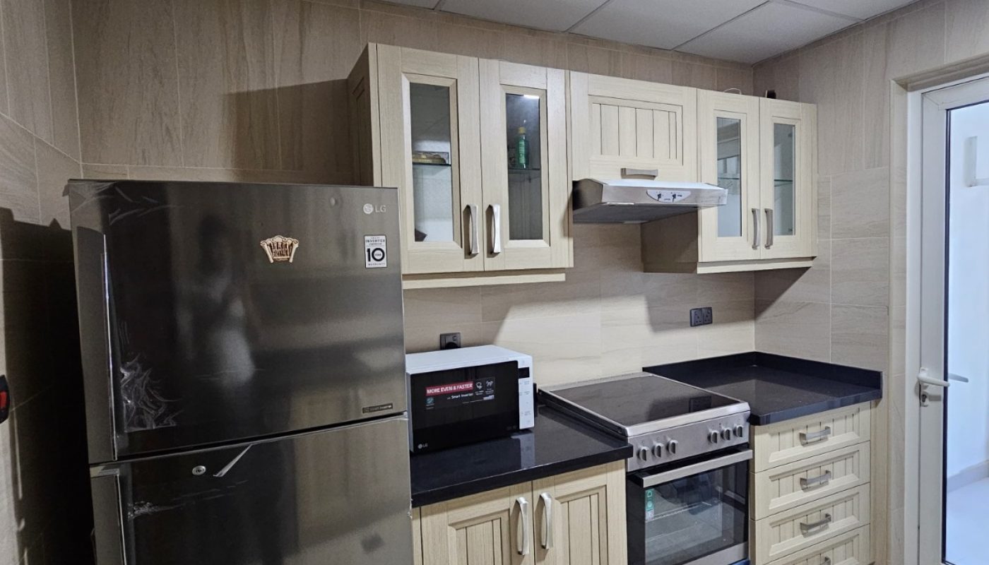 Modern kitchen with light wood cabinets, stainless steel refrigerator, microwave, stove, and oven, black countertops, and beige tiled floor.