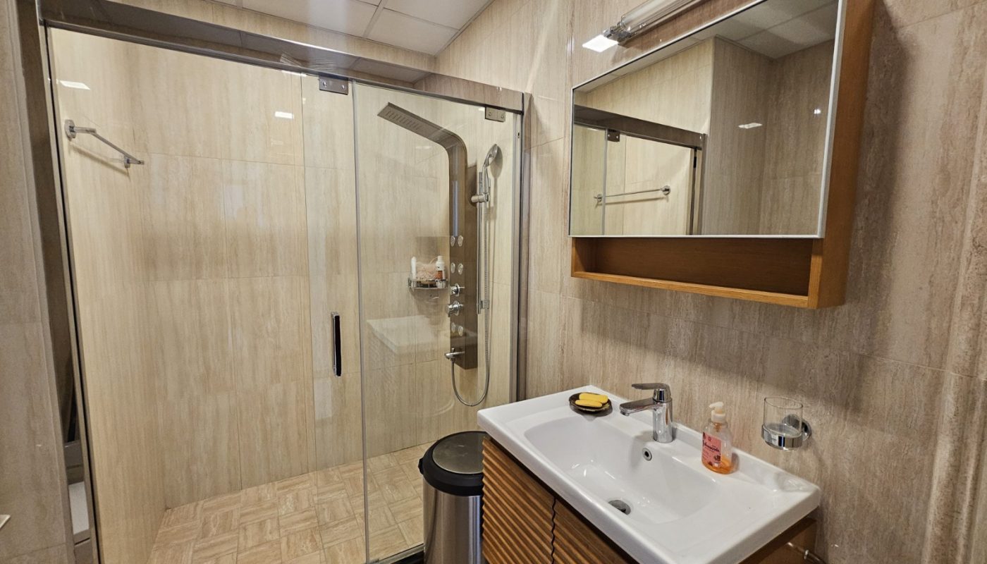 Modern bathroom with a glass-enclosed shower, wall-mounted sink with cabinet, mirror cabinet above, and a trash bin beside the sink.