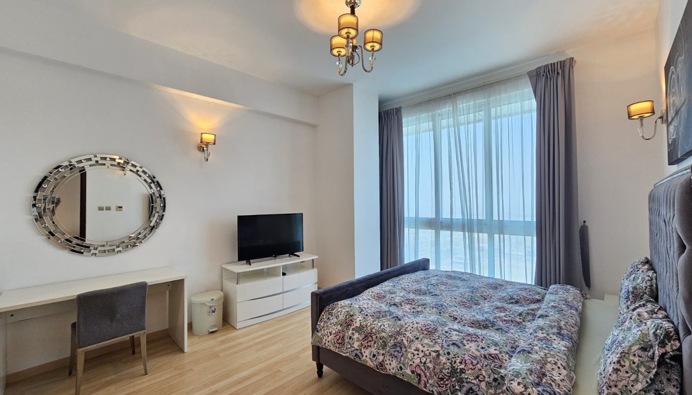 A modern bedroom with a double bed, floral bedding, a TV on a white dresser, a desk with a chair, wall mirrors, and large windows with curtains letting in natural light.