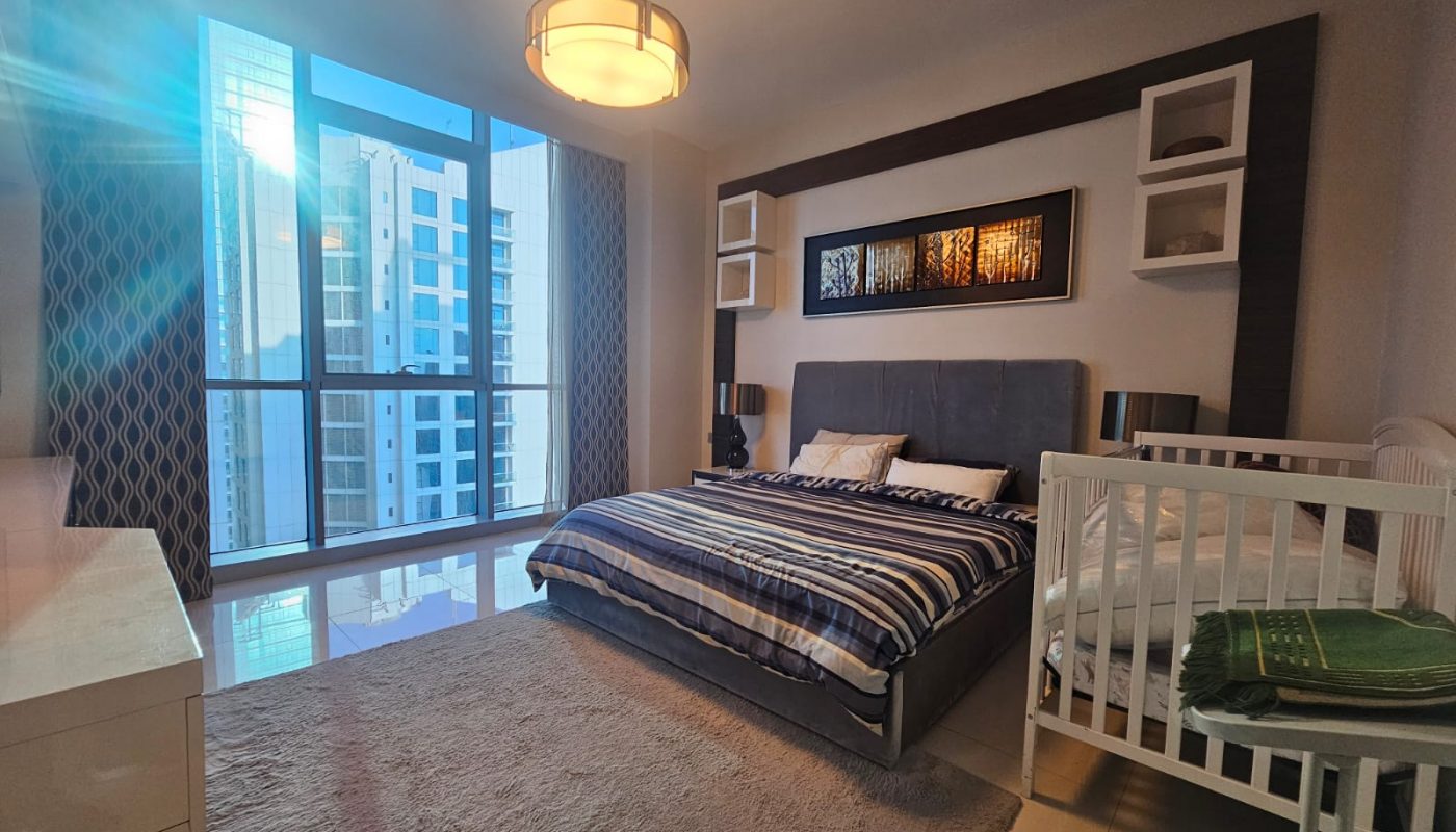 A modern bedroom with a large bed, a crib, a dresser, a rug, and floor-to-ceiling windows showing tall buildings outside. Sunlight enters from the left side.