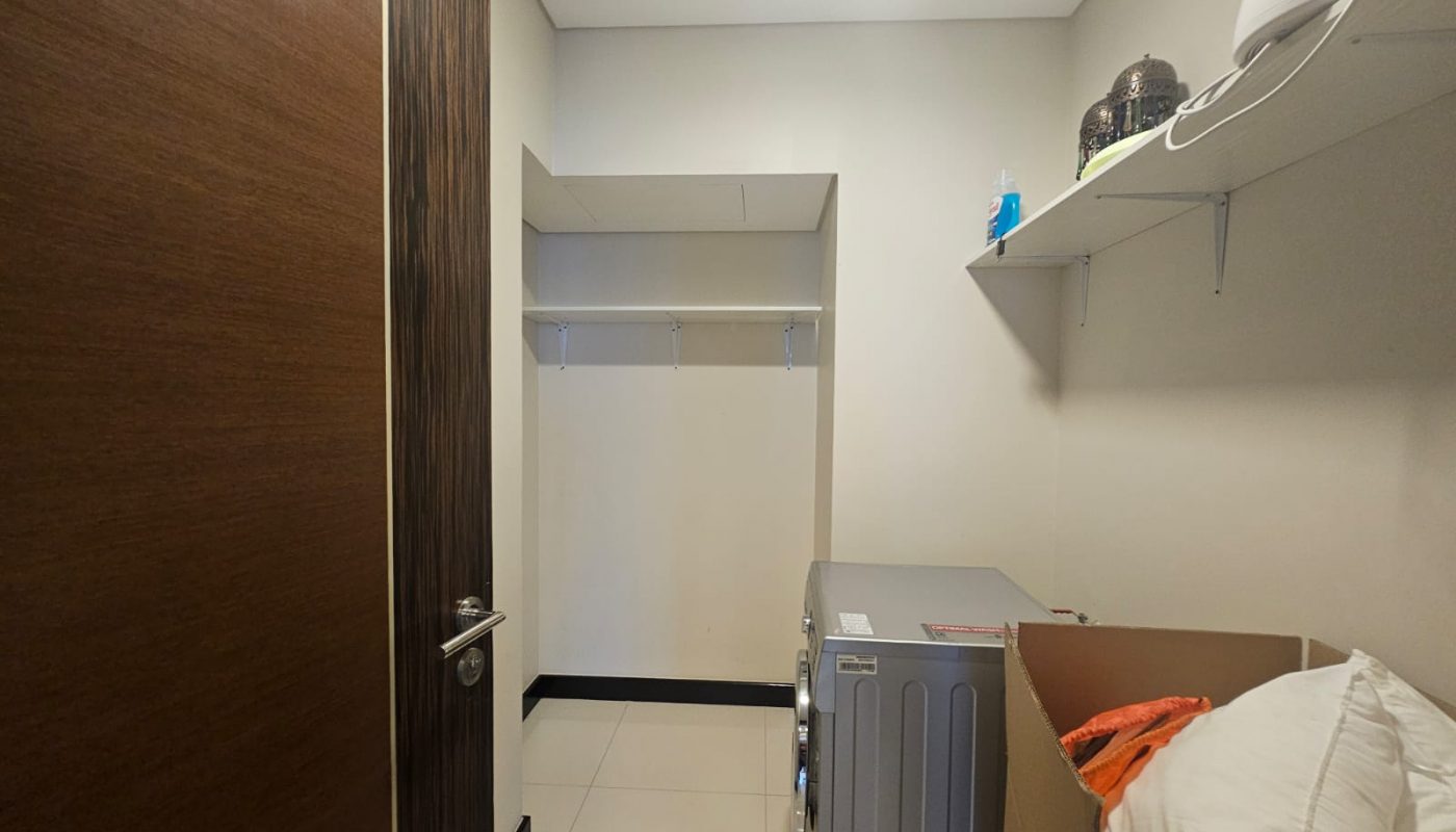 A small laundry room with a washing machine, a cardboard box with pillows, a wall shelf holding containers, and an empty open closet with hangers.