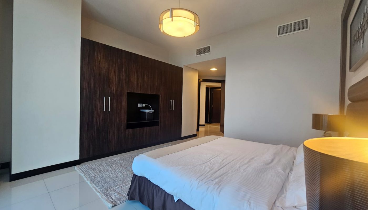 A modern bedroom with a large bed, built-in dark wood wardrobe, tiled floor, ceiling light fixture, and neutral-colored walls.
