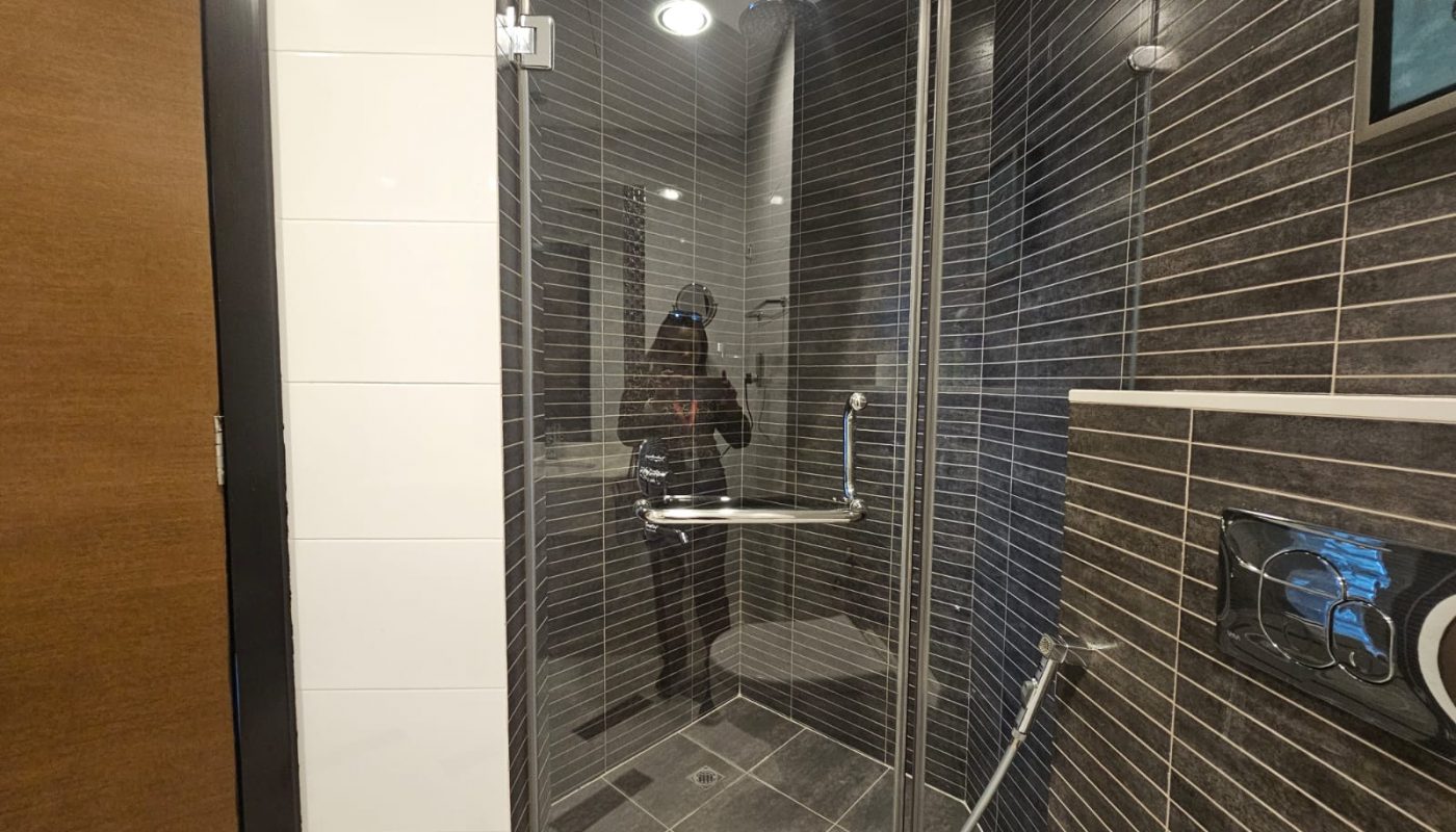 A person is reflected in the glass door of a modern shower with dark tiles, a rainfall showerhead, and a handheld shower attachment.
