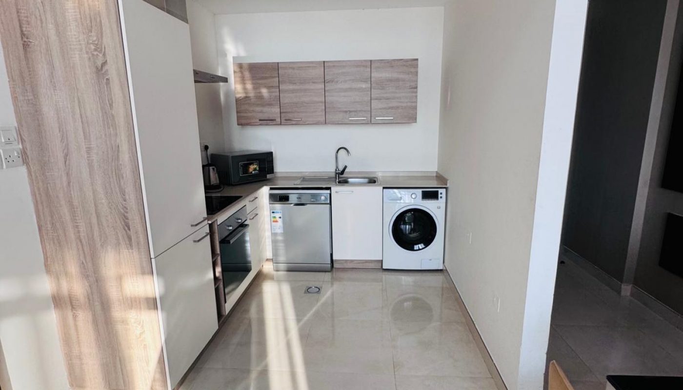 Modern kitchen with light wood cabinets, built-in oven, microwave, dishwasher, and washing machine; sunlit tile floor and a chair visible in the foreground.