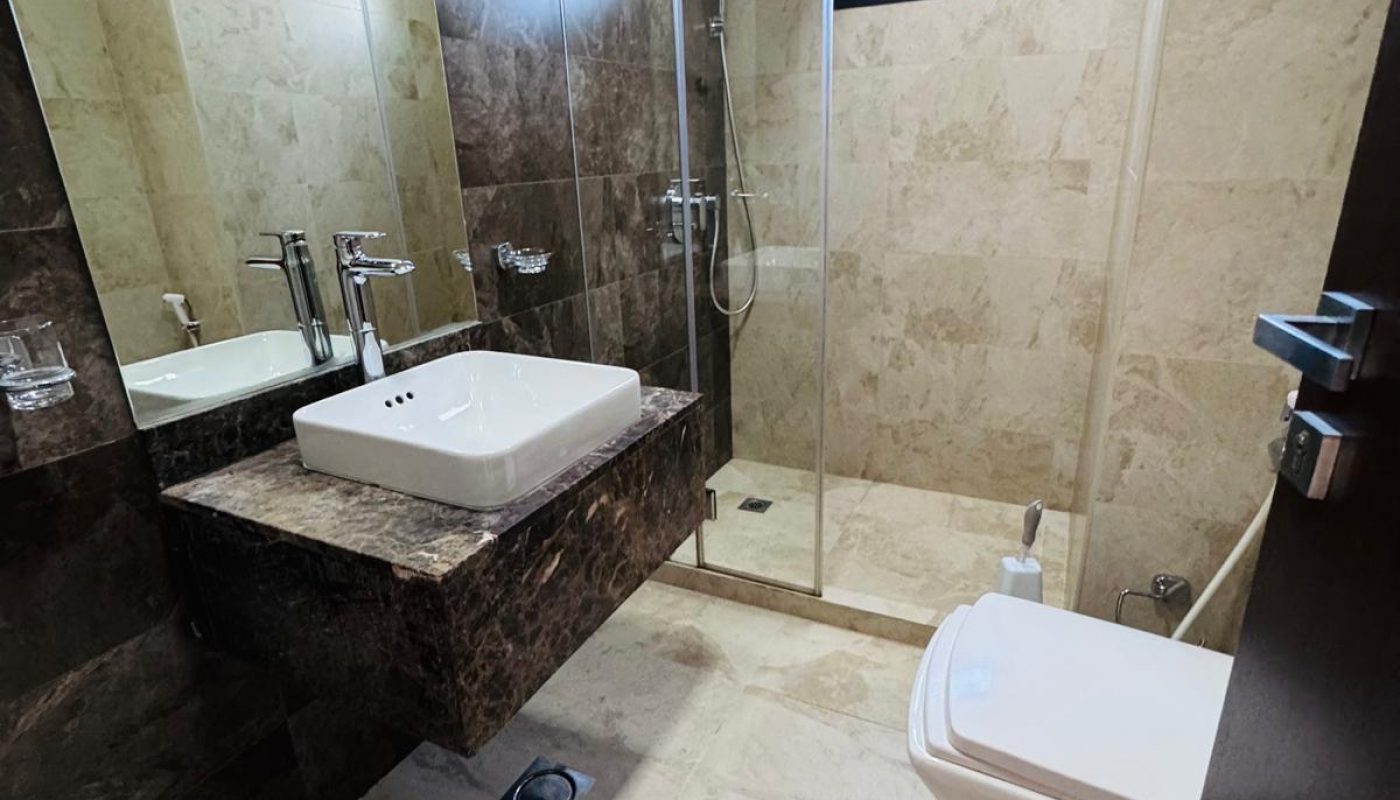 Modern bathroom with beige tiles, wall-mounted sink, glass-enclosed shower, toilet, and a window for natural light.