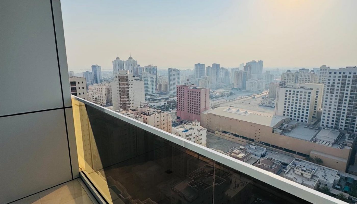 A high-rise balcony with a glass railing overlooks a cityscape of tall buildings under a clear sky with the sun visible.