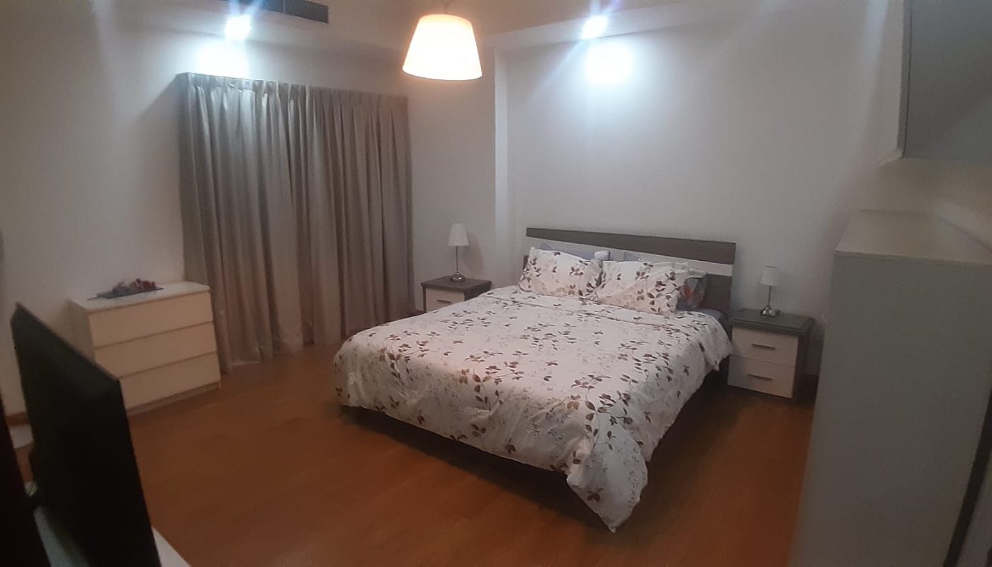 A bedroom with a double bed covered in a floral-patterned duvet, two bedside tables with lamps, a chest of drawers, and closed beige curtains over a window.