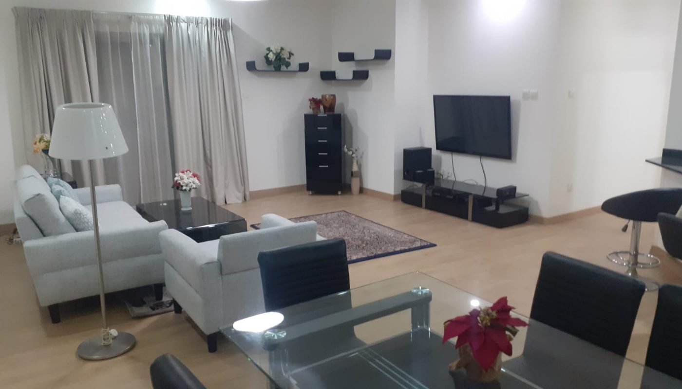 Modern living room with light-colored sofas, glass coffee and dining tables, TV mounted on the wall, shelves with decor, and a floor lamp. Curtains cover a large window or balcony door.