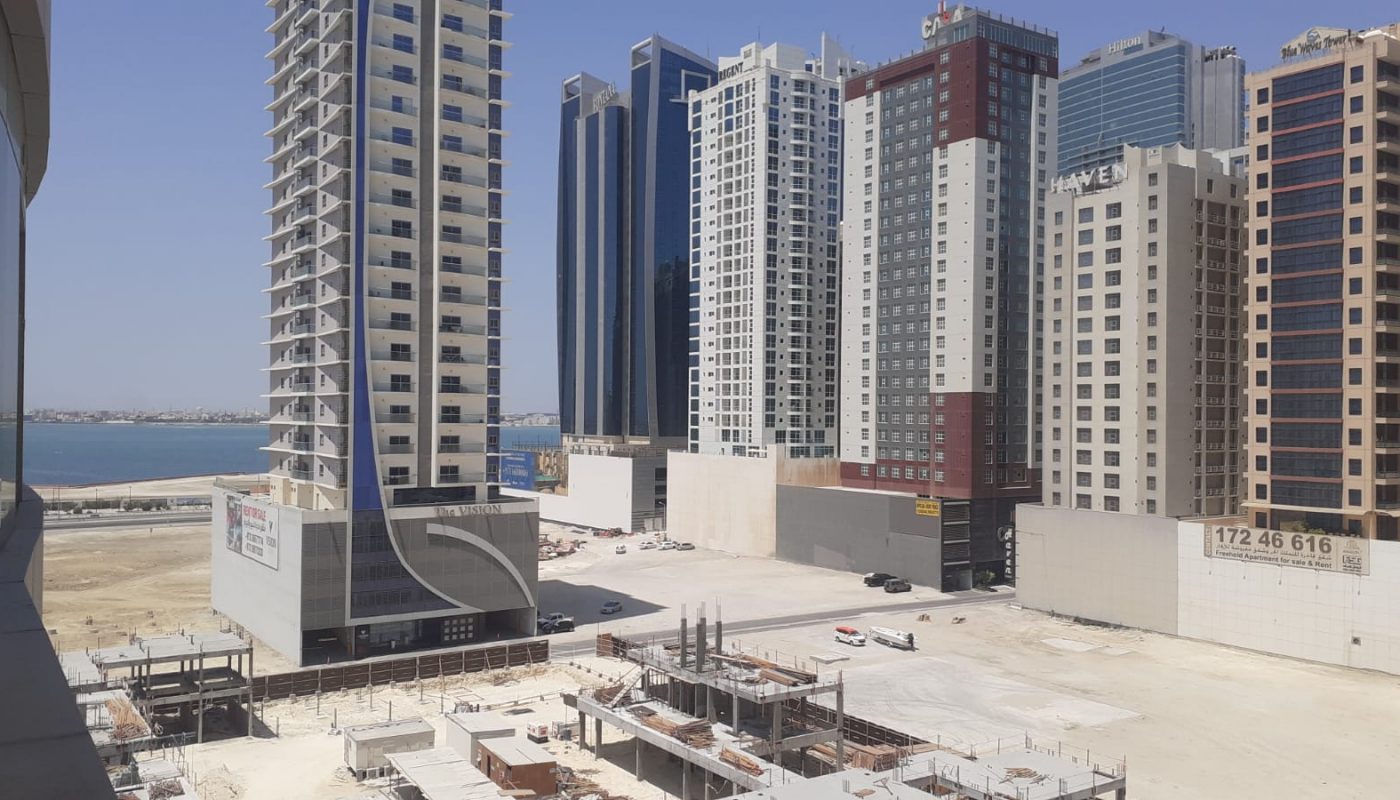 High-rise buildings and construction sites stand near a waterfront under a clear blue sky, with a few parked cars and unfinished structures visible.