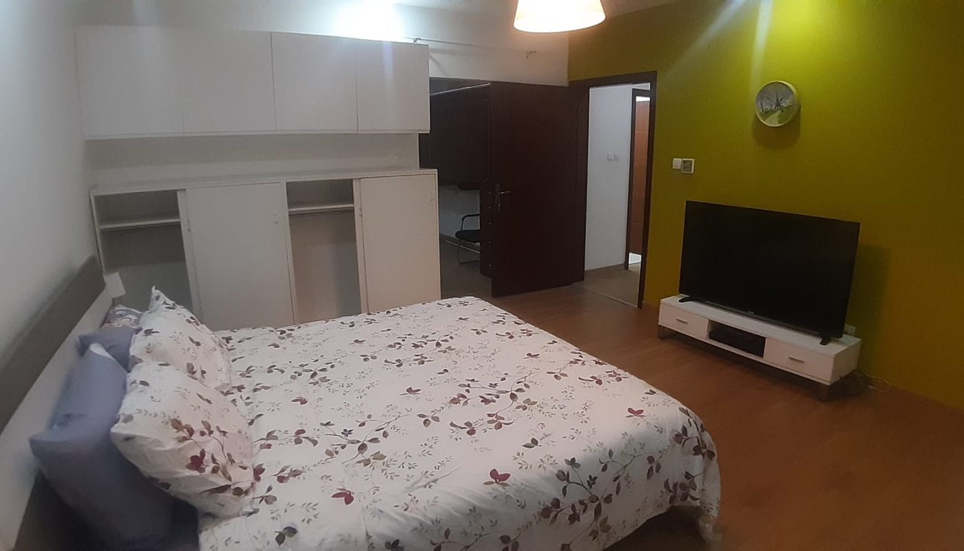 A bedroom with a floral bedspread, wall-mounted cabinets, open closet, TV on a stand, green accent wall, and visible open doors.
