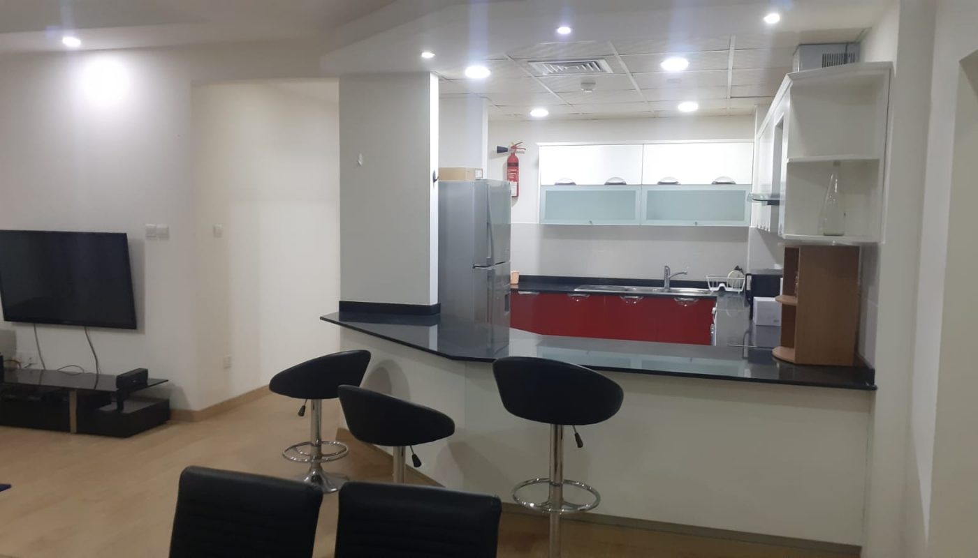Modern open-plan kitchen with black countertops, bar seating, white cabinets, and stainless steel appliances, adjacent to a living area with a wall-mounted TV.