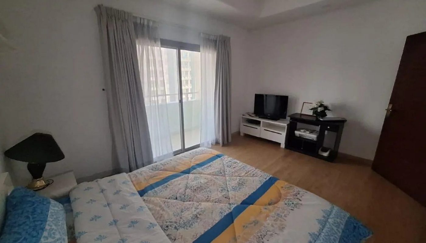 A bedroom with a bed, a TV on a stand, a small table with a plant, a lamp, and a sliding glass door with curtains leading to a balcony.