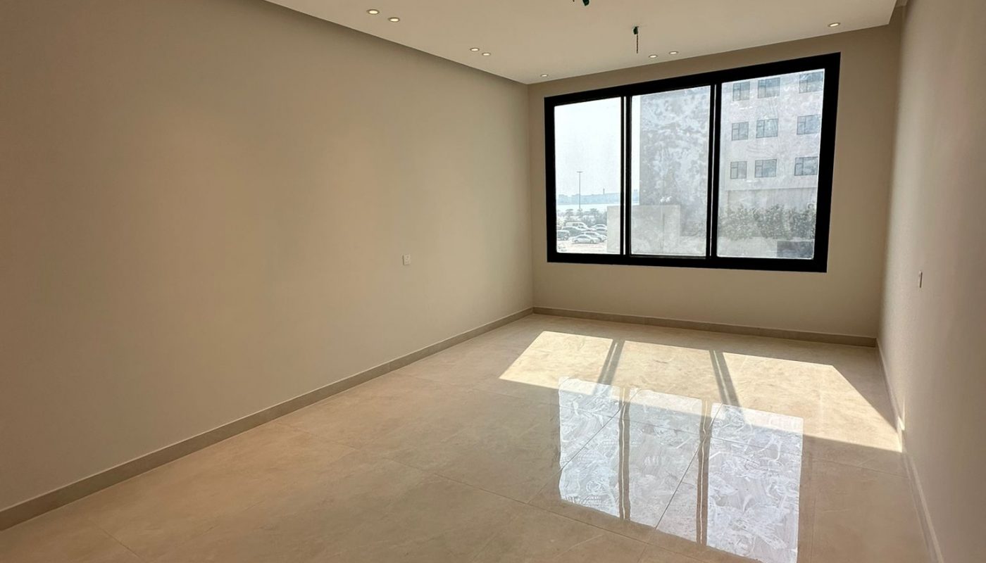 An empty, unfurnished room with beige tile floors, light-colored walls, ceiling lights, and a large window letting in sunlight.