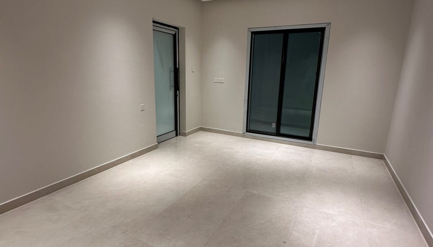 Empty, unfurnished room with light-colored tiled floor, beige walls, recessed ceiling lights, sliding glass door, and an open doorway on the left.