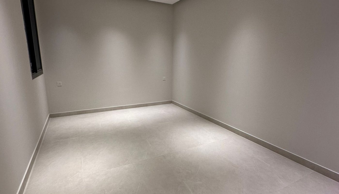 Empty room with beige tiled floor, off-white walls, recessed ceiling lights, and a single window on the left wall. No furniture or decorations are present.