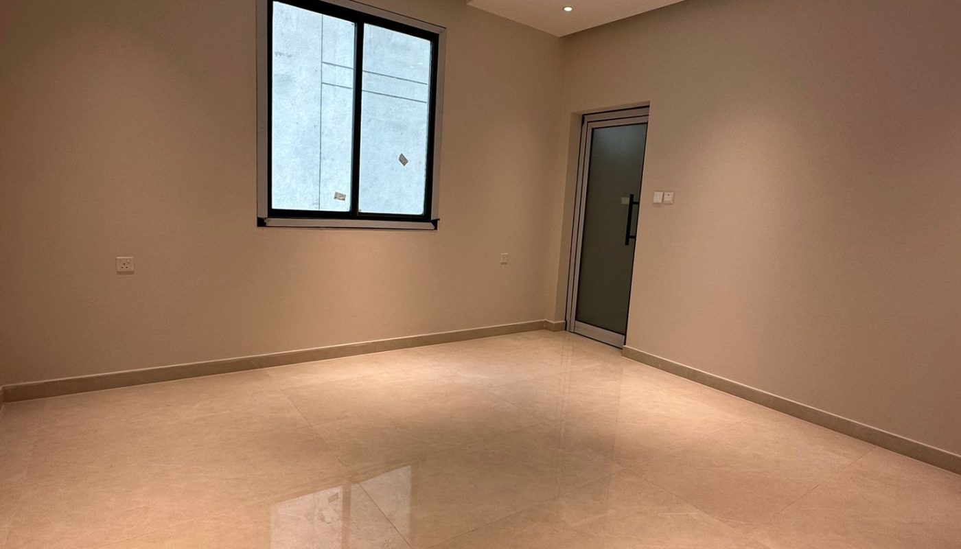 Empty room with beige tiled floor, cream-colored walls, recessed ceiling lights, a window with black frame, and a closed door. A wire hangs from the ceiling.