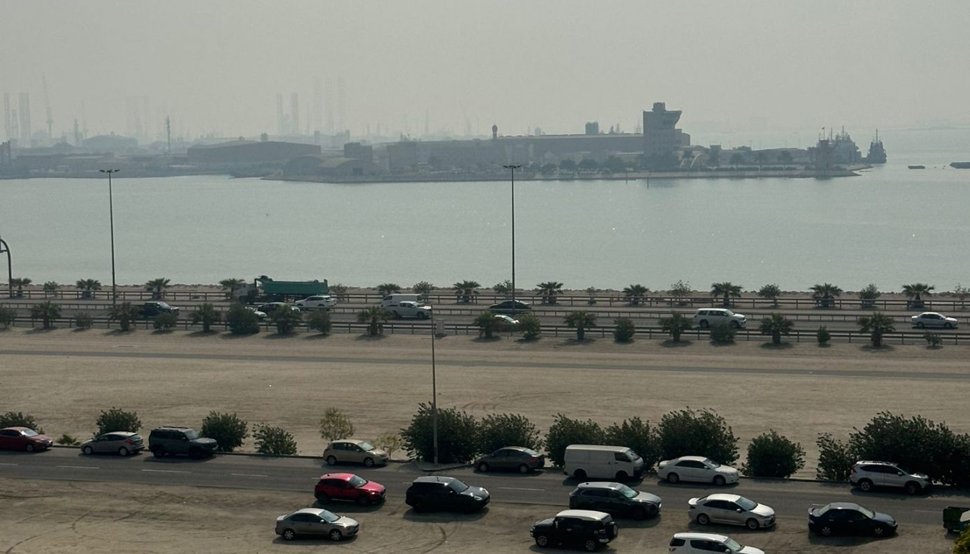 A dusty parking lot with scattered cars in the foreground, a road lined with trees, and a hazy waterfront with docked ships in the background.