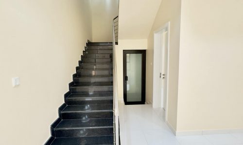 Interior of a luxury villa in Maqabah Saraya 1, showing a staircase with black steps leading up to the next floor, alongside a well-lit, empty hallway with white walls and