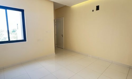 A luxury 4 bedrooms villa for sale in Maqabah, featuring an empty room with white tiled flooring, pale yellow walls, recessed ceiling lights, and a window on the left side.