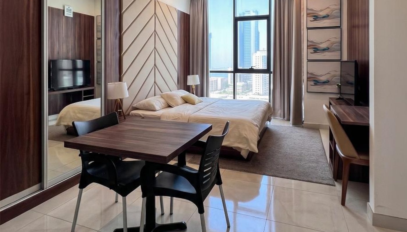 Modern studio apartment with a dining table, bed, wall-mounted TV, large windows, and city view, featuring minimalist decor and tiled flooring.