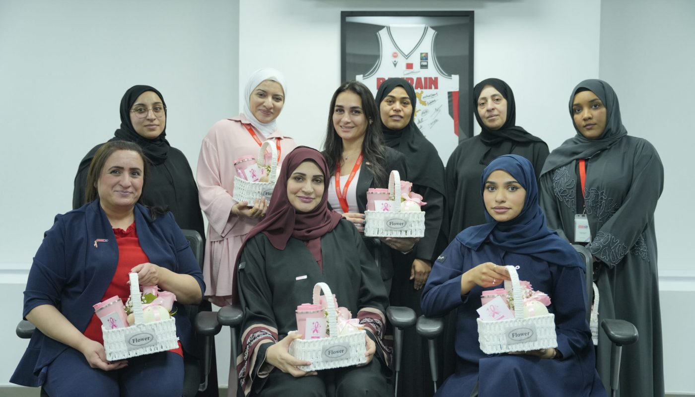 Nine women pose indoors at an October awareness event, holding baskets with pink ribbons. A framed sports jersey is displayed on the wall behind them, highlighting House Me’s commitment to community causes.