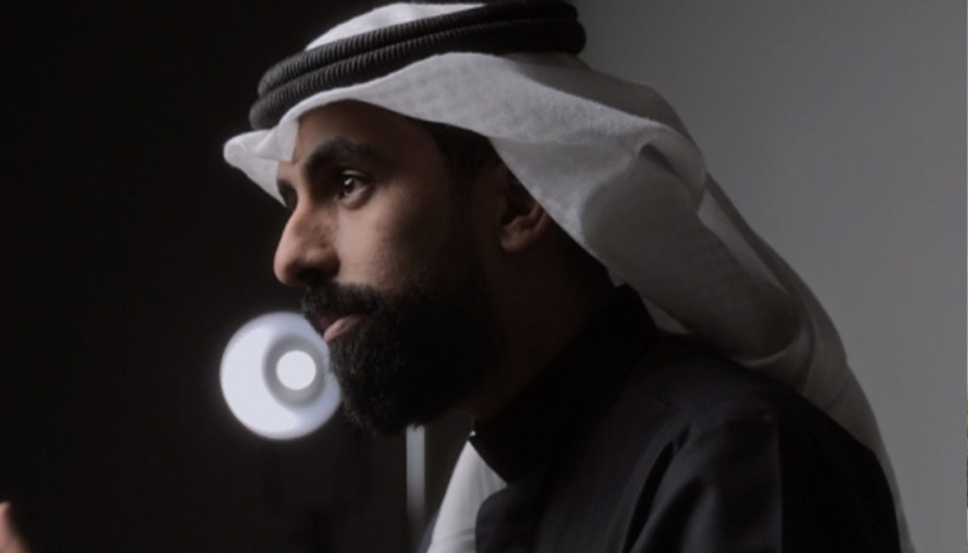 A person with a beard, symbolizing new beginnings, dons a black robe and a white keffiyeh, bathed in the glow of a round light.