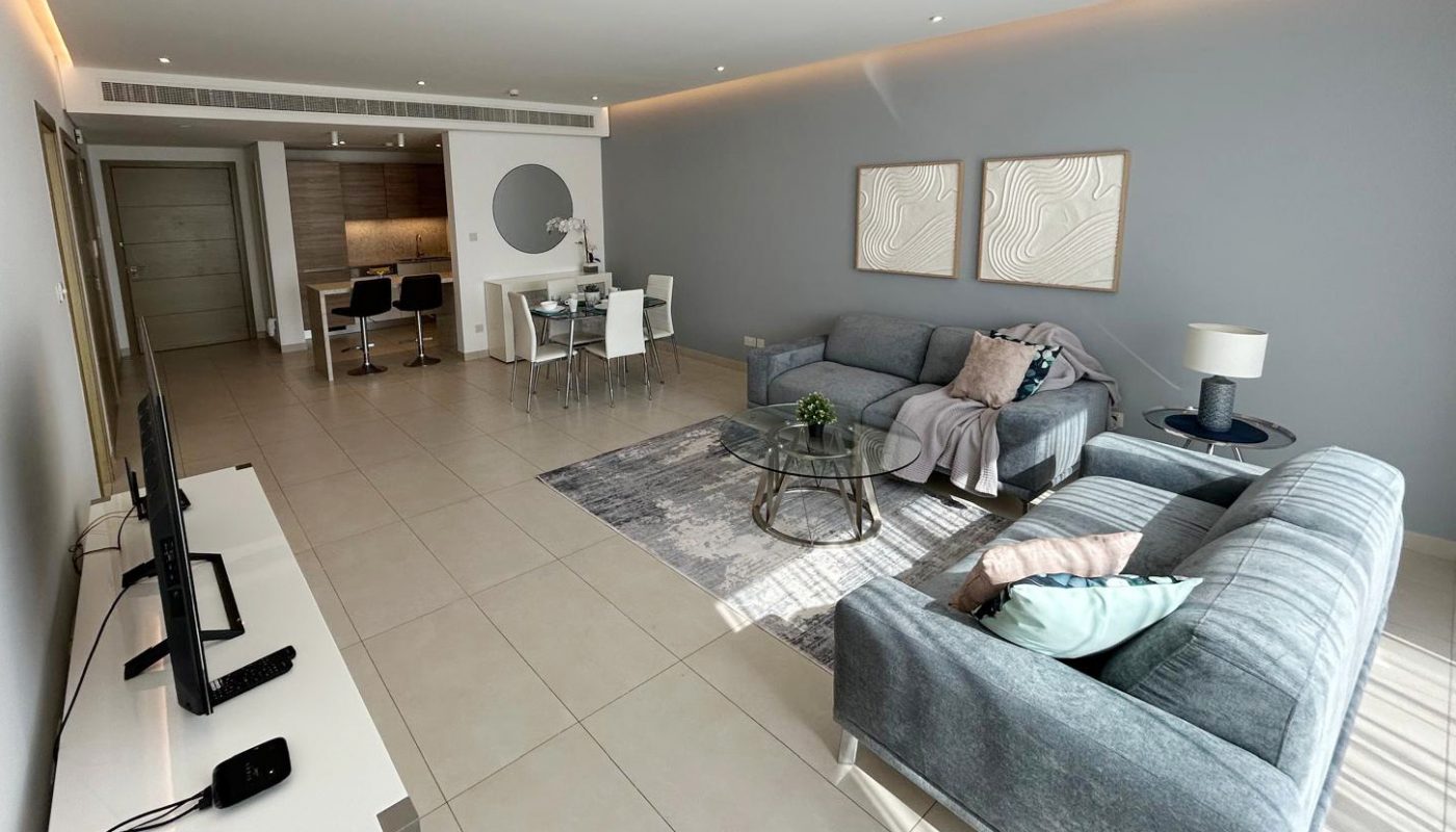 A modern living and dining area with two gray sofas, a TV on a white stand, a dining table with six chairs, and minimalistic decor on light tiled floors.