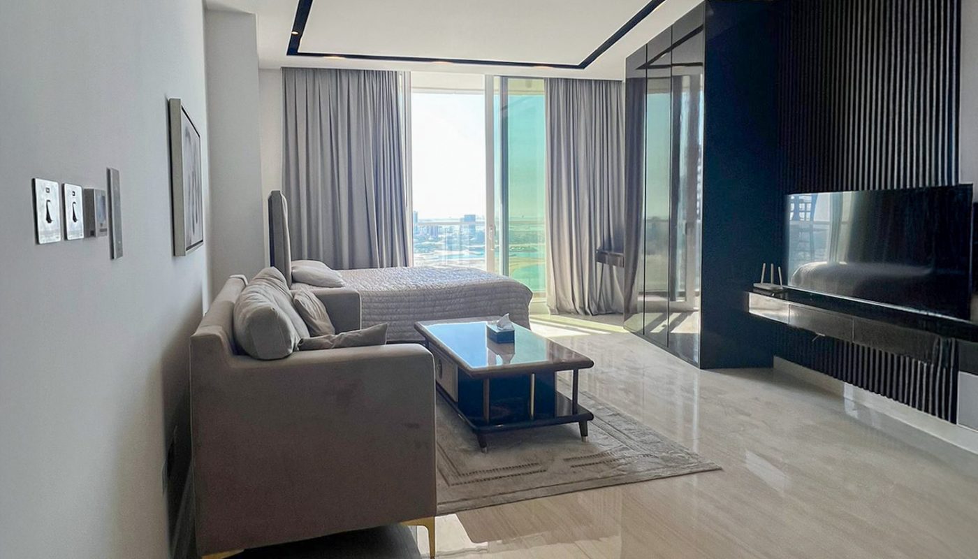 Modern living room with beige tiled floors, a gray sofa, coffee table, large windows with curtains, and a wall-mounted TV. Natural light fills the space.