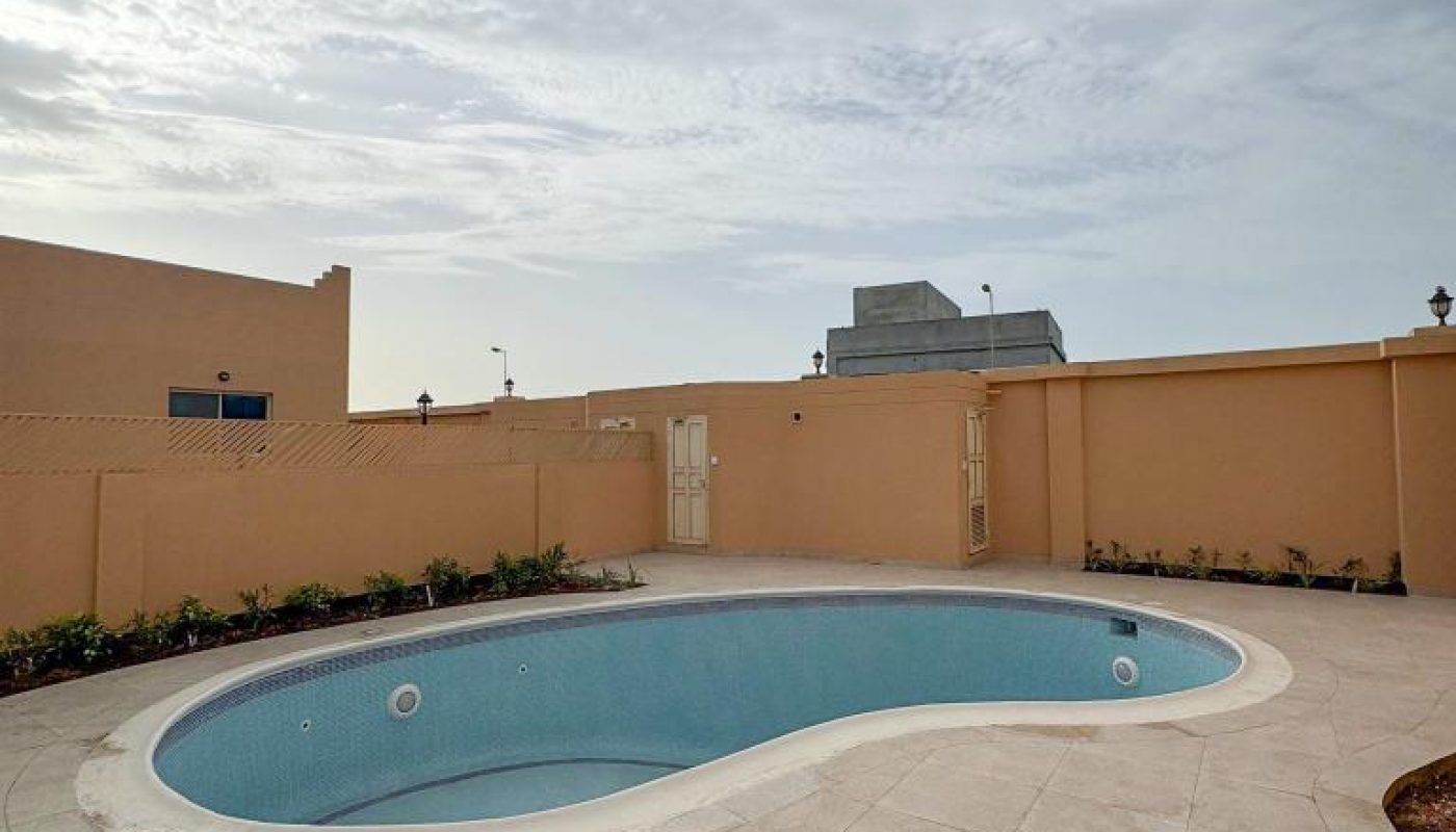 An empty kidney-shaped swimming pool with beige tile decking, surrounded by a tan wall, under a cloudy sky.