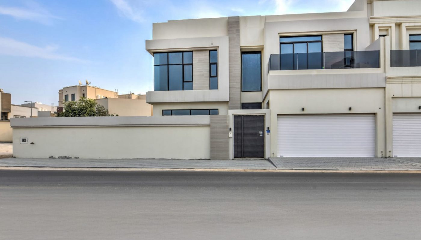 A modern two-story house with large windows, white exterior walls, and two garage doors, viewed from across an empty street under a partly cloudy sky.