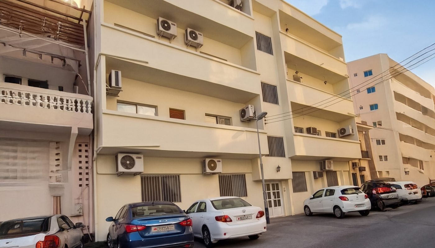 Four-story apartment building with multiple balconies and air conditioning units, parked cars in front, and neighboring buildings visible under a clear blue sky.