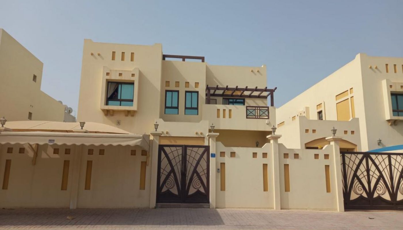 A two-story beige house with modern geometric architectural features, blue-tinted windows, a gated driveway, and a covered carport on a sunny day.