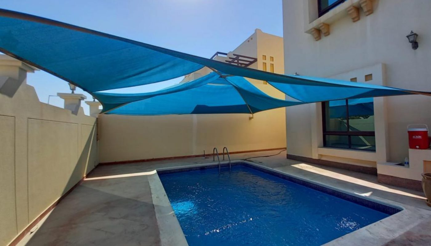 Rectangular outdoor swimming pool beside a beige building, shaded by large blue fabric canopies, with sunlight above.