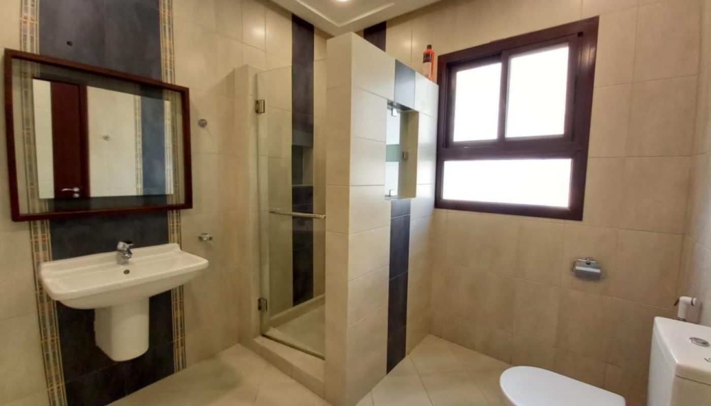 Modern bathroom with beige tiled walls, a glass-enclosed shower, a wall-mounted sink with mirror, a toilet, and a large window.